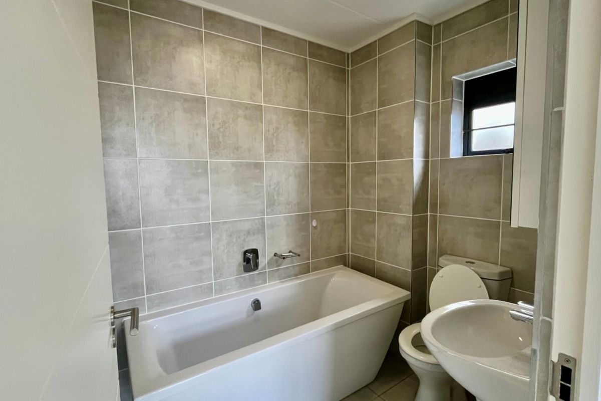 A clean, modern bathroom featuring a white bathtub, pedestal sink, toilet, and neutral-toned wall tiling.