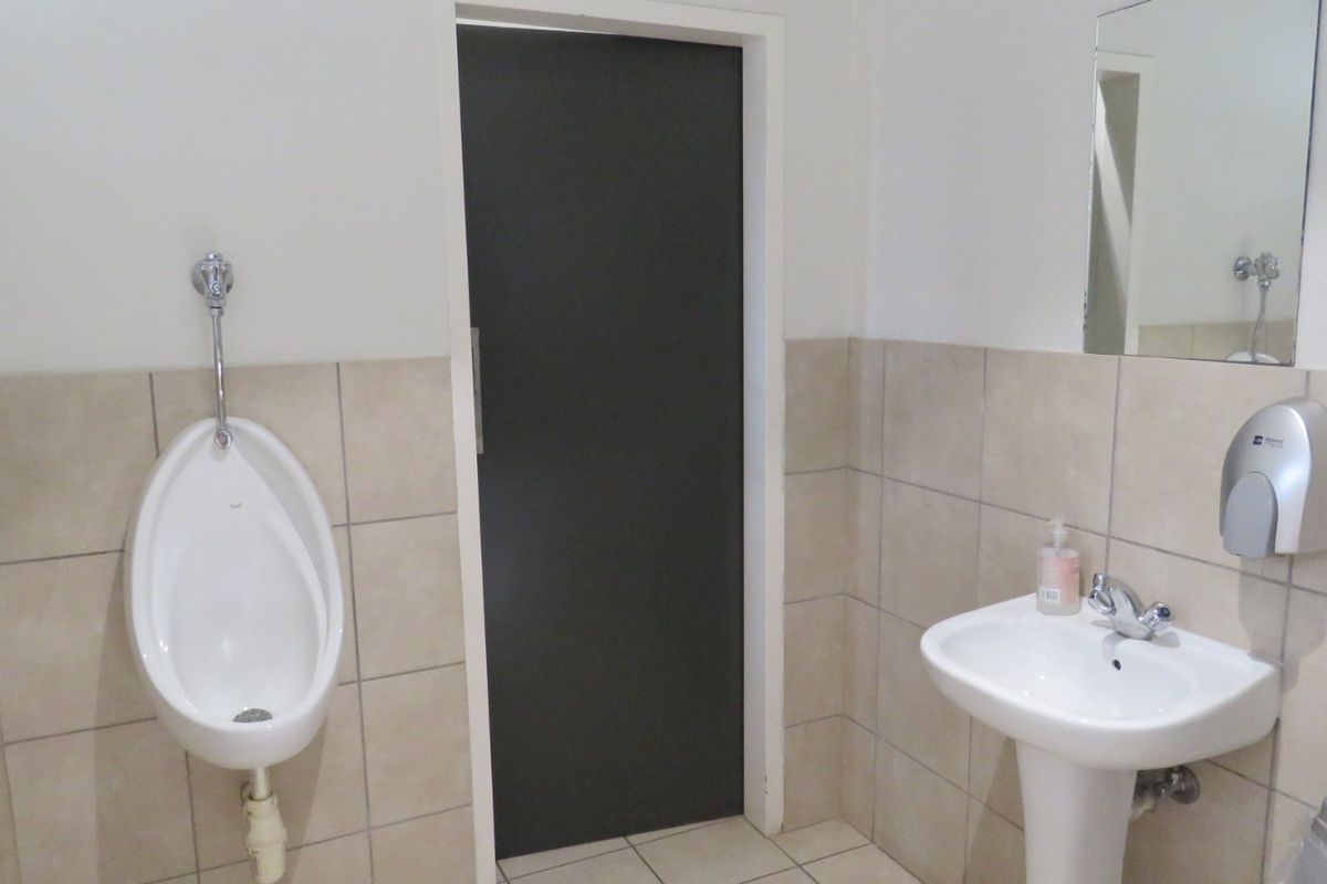 A functional commercial bathroom featuring a wall-mounted urinal, pedestal basin, tiled wall finishes, and a mirror.
