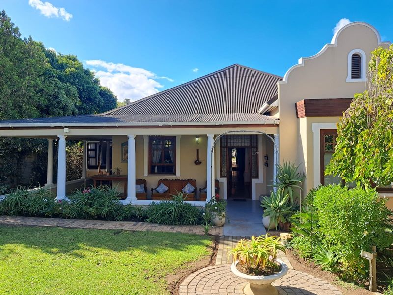 Charming front elevation featuring a covered veranda with classical columns, a corrugated metal roof, and a well-maintained garden with a paved walkway.