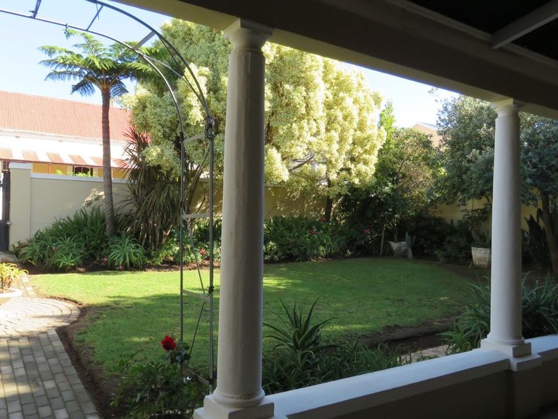 A covered patio overlooking a well-maintained garden with a paved walkway, lush lawn, and secure boundary wall with a metal gate.