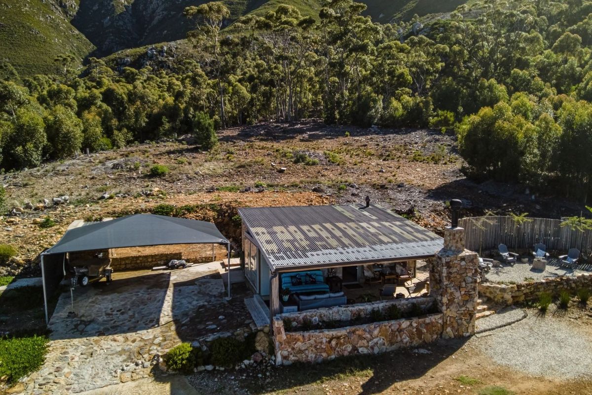 Aerial view of a small holding property featuring a dwelling with a corrugated roof, stone chimney, and an attached carport set against a mountain backdrop.