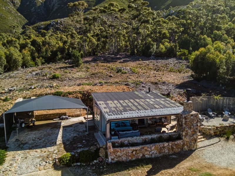 Aerial view of a small holding property featuring a dwelling with a corrugated roof, stone chimney, and an attached carport set against a mountain backdrop.
