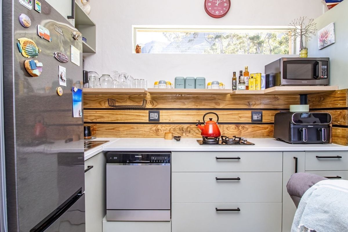 Compact modern kitchen featuring light grey cabinetry, a wooden splashback, integrated dishwasher, gas hob, and floating shelving under a horizontal clerestory window.