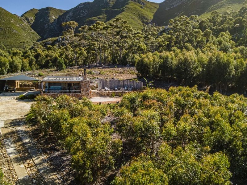 Aerial view of a rustic stone-clad small holding dwelling nestled against a mountain backdrop, featuring a covered patio and natural landscape surroundings.
