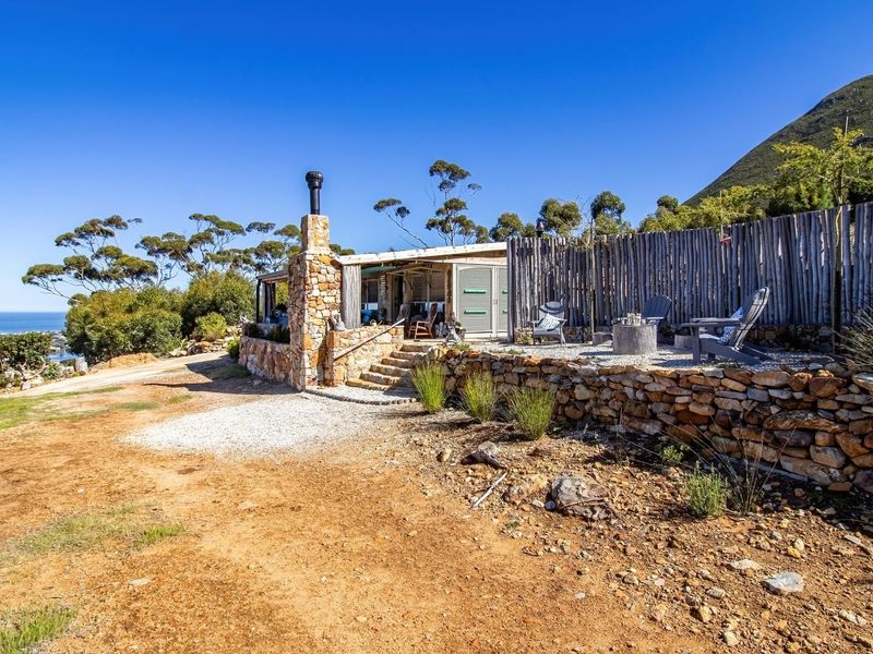 A rustic rear exterior view featuring a stone retaining wall, stone chimney, and gravel patio area with scenic coastal and mountain views.