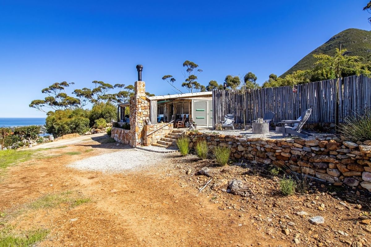 A rustic rear exterior view featuring a stone retaining wall, stone chimney, and gravel patio area with scenic coastal and mountain views.