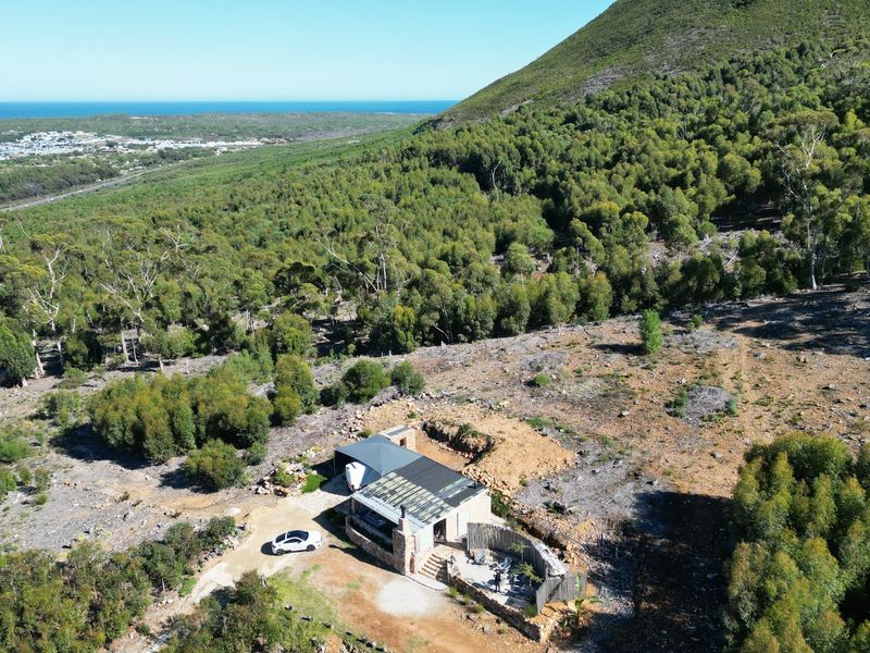 Aerial view of a small holding property nestled against a forested hillside, featuring a stone-clad dwelling with a metal roof and scenic coastal views.