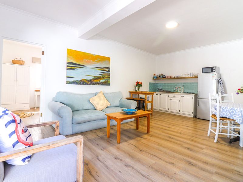 Open-plan living area featuring light wood-look laminate flooring, a compact kitchen with tiled backsplash, and neutral white walls.