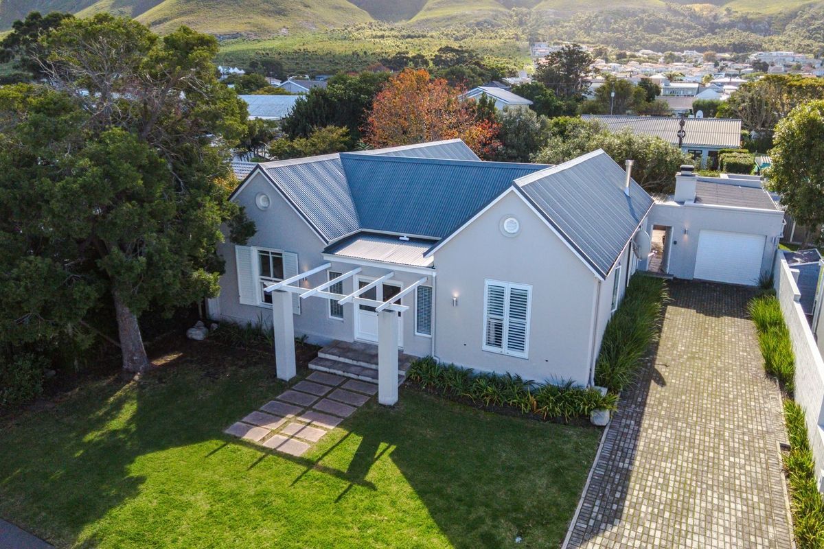 Aerial view of a well-maintained single-story house featuring a metal roof, front pergola, paved driveway, and attached garage set in a landscaped garden.