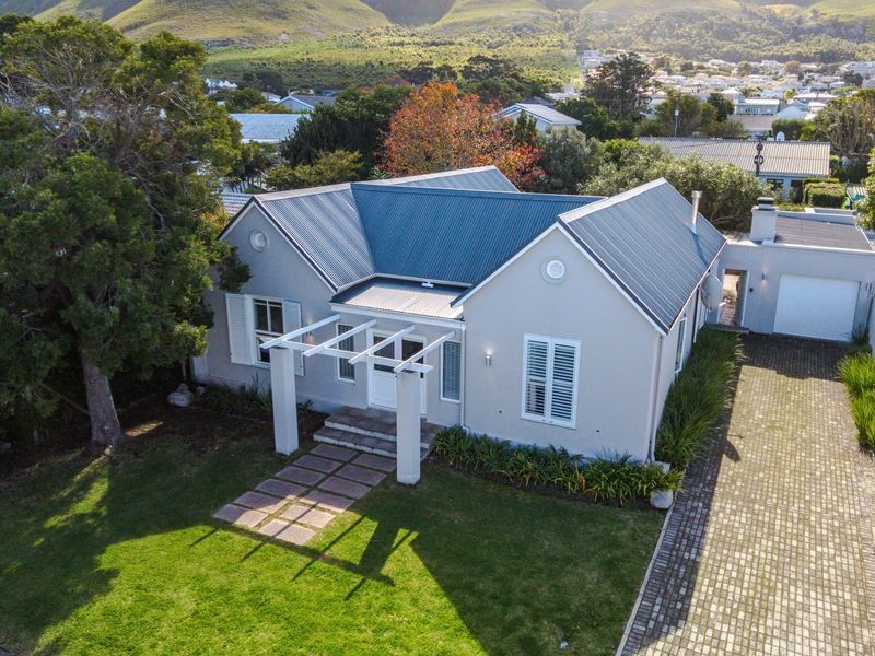 Aerial view of a well-maintained single-story house featuring a metal roof, front pergola, paved driveway, and attached garage set in a landscaped garden.