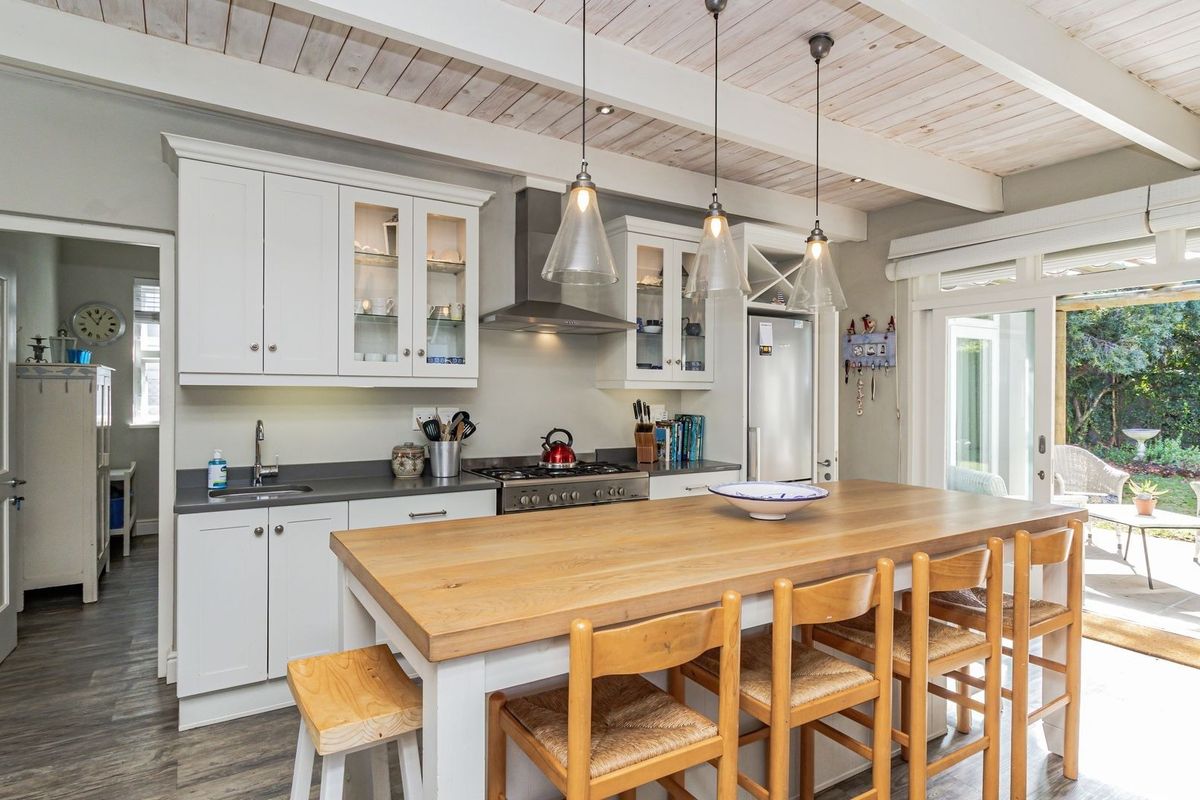 A bright, modern kitchen featuring shaker-style cabinetry, stone countertops, a stainless steel gas range, and exposed timber beam ceilings, opening directly to the garden.