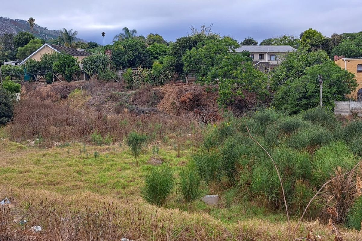 A view of a vacant residential plot featuring natural vegetation, uneven terrain, and surrounding established residential properties.