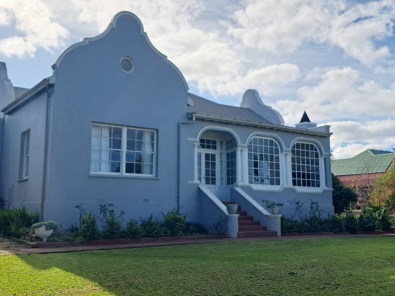 A charming Cape Dutch style house featuring a distinctive curved gable, arched windows, and a welcoming columned entrance set within a well-maintained garden.