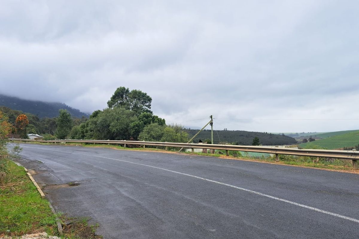 A view of the asphalt road and guardrail bordering the vacant residential land, featuring surrounding greenery and distant hills.
