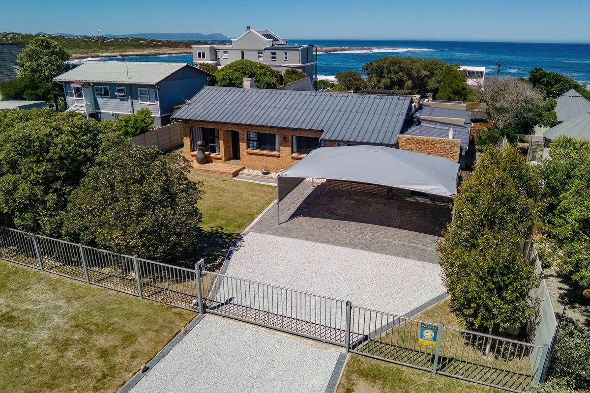 Aerial view of a coastal brick house featuring a metal roof, paved driveway, carport, and secure perimeter fencing with ocean views.