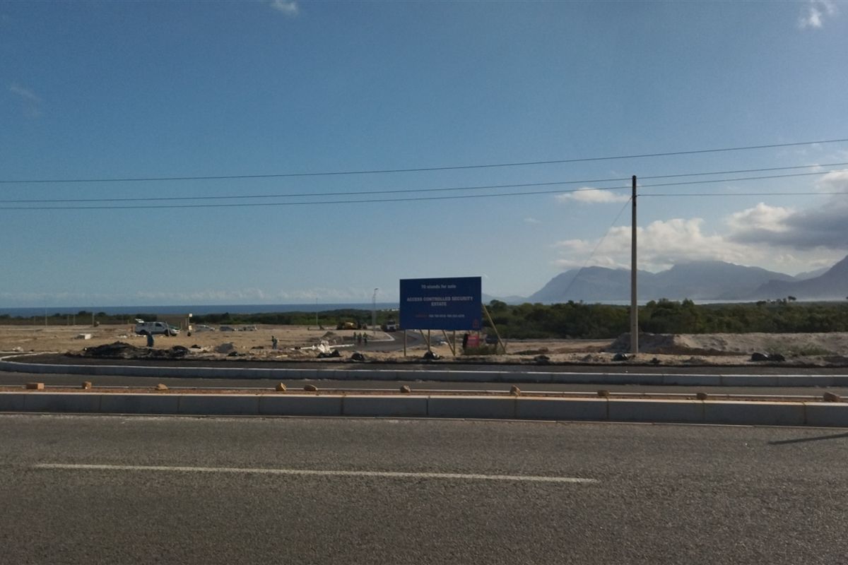 A street-level view of a vacant commercial development site featuring paved road infrastructure, curbing, and prominent signage against a mountain and coastal backdrop.