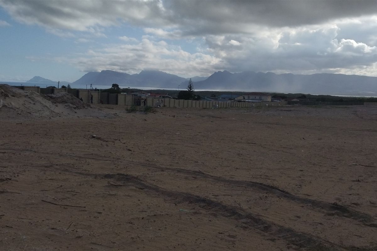 A wide view of a vacant commercial land plot featuring sandy terrain and a perimeter wall with distant mountain views.