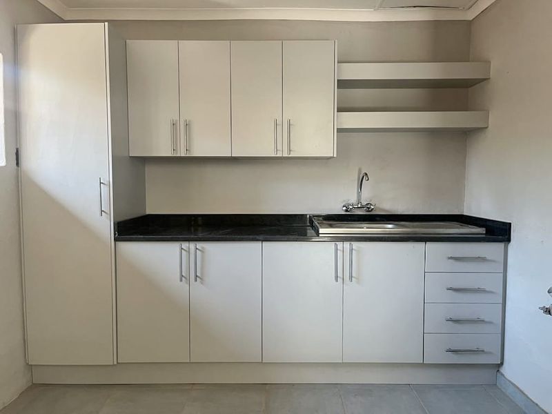 A clean, functional kitchen area featuring modern white cabinetry, dark laminate countertops, a stainless steel sink, and convenient floating wall shelves.