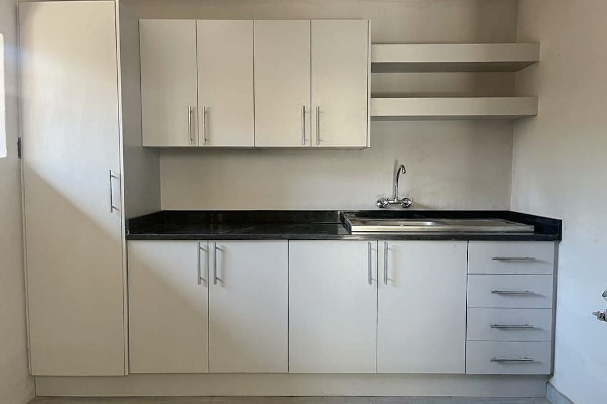 A clean, functional kitchen area featuring modern white cabinetry, dark laminate countertops, a stainless steel sink, and convenient floating wall shelves.