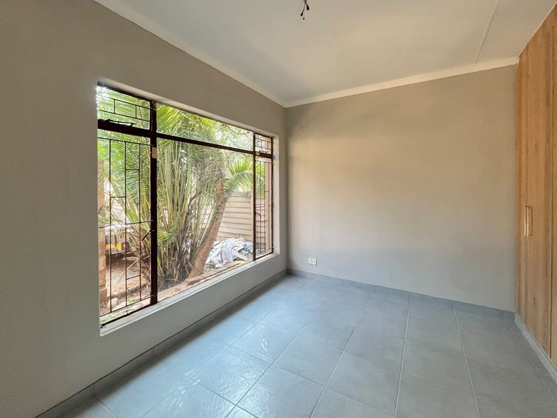 A bright, empty bedroom featuring tiled flooring, built-in wooden cupboards, and a large window with security bars overlooking the garden.