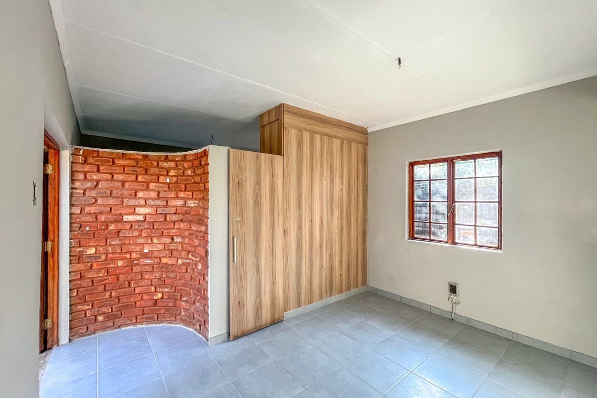 A bright, empty bedroom featuring a unique curved exposed brick wall, built-in wooden wardrobe, tiled flooring, and a classic wooden window frame.