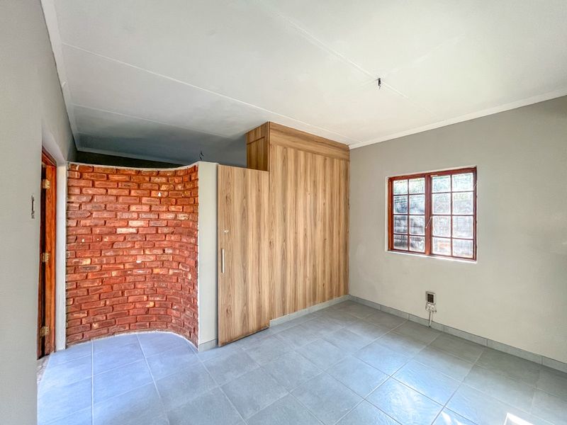 A bright, empty bedroom featuring a unique curved exposed brick wall, built-in wooden wardrobe, tiled flooring, and a classic wooden window frame.