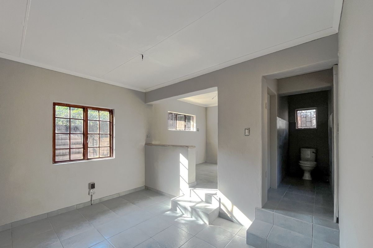 An empty living area featuring tiled flooring, timber-framed windows, and an adjacent bathroom with a toilet, requiring minor cosmetic repairs.