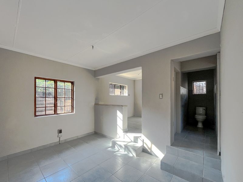 An empty living area featuring tiled flooring, timber-framed windows, and an adjacent bathroom with a toilet, requiring minor cosmetic repairs.