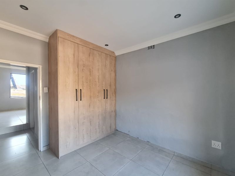 A clean, empty bedroom featuring light wood-finish built-in wardrobes, neutral grey walls, tiled flooring, and recessed ceiling lighting.