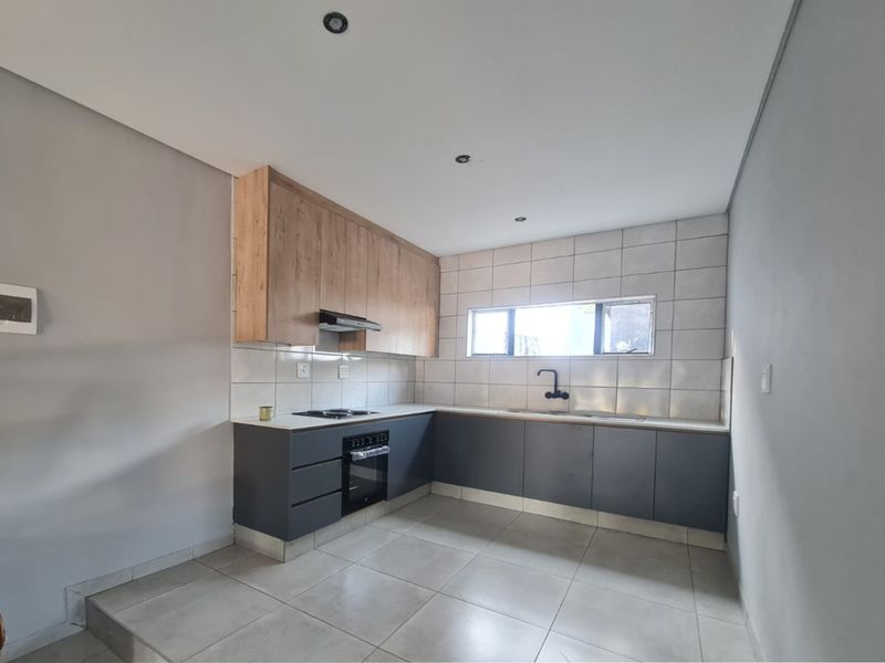 Modern kitchen featuring grey lower cabinetry, wood-look upper cabinets, tiled backsplash, built-in oven and hob, and neutral floor tiles.