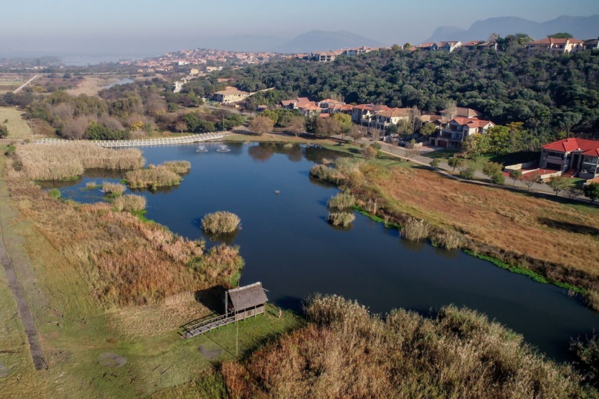 Aerial view of the estate featuring a large lake, landscaped surroundings, a thatched gazebo, and a wooden walkway.