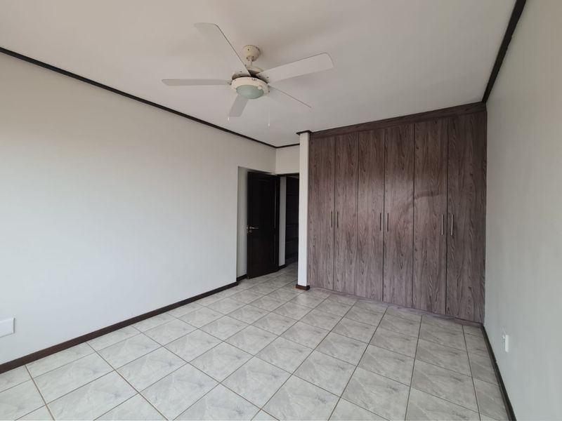 A spacious, empty bedroom featuring neutral tiled flooring, a ceiling fan, and extensive built-in wooden cupboards for ample storage.