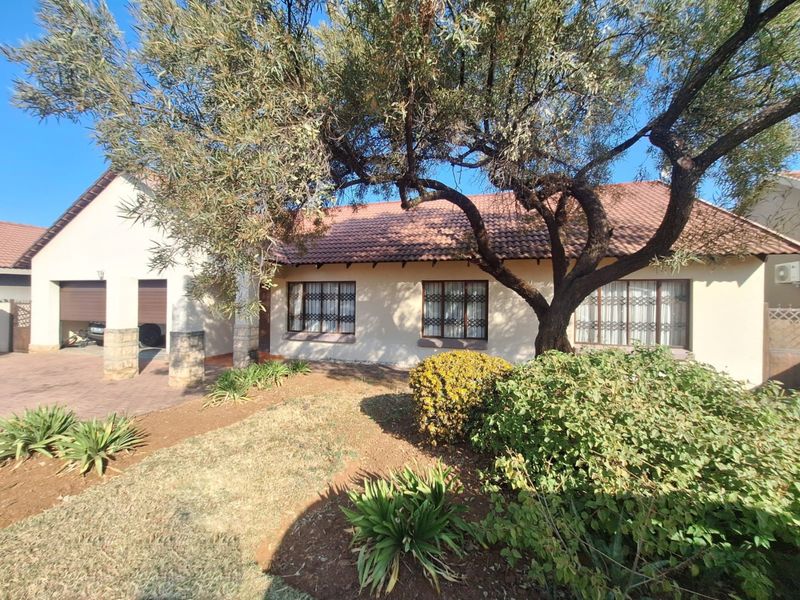 Front elevation of a single-story house featuring a tiled roof, double garage, paved driveway, and established garden with mature trees.