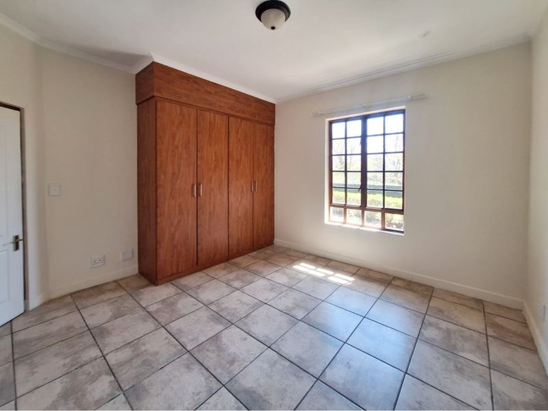 A bright, empty bedroom featuring tiled flooring, a built-in wooden wardrobe, and a grid-pattern window providing natural light.