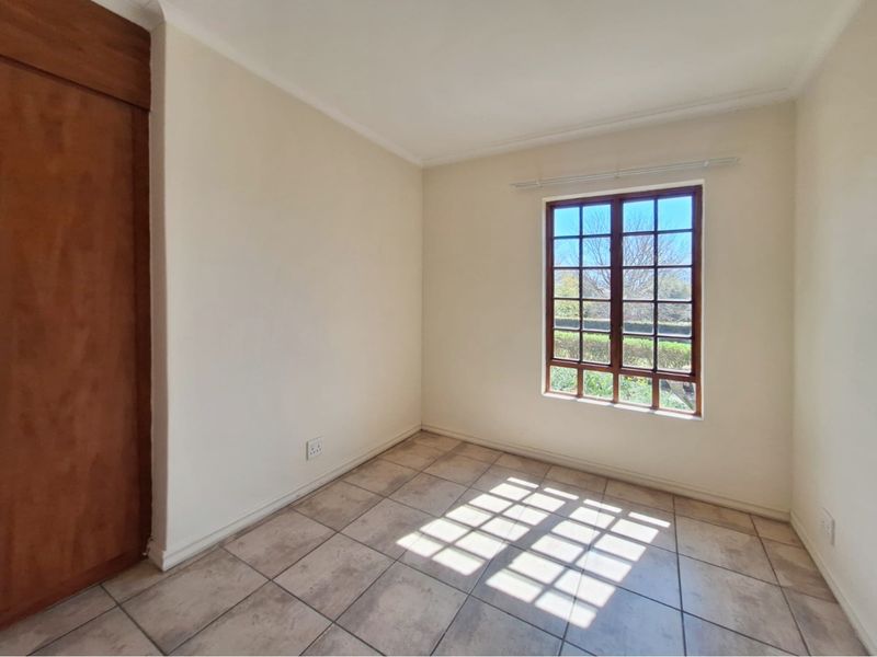 A bright, empty bedroom featuring tiled flooring, a built-in wooden cupboard, and a window with a garden view.
