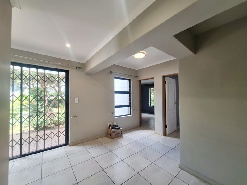 An empty, neutrally painted living area featuring tiled flooring, a sliding door with a security gate, and access to adjacent rooms.