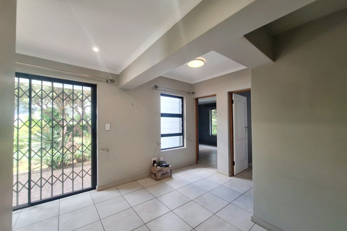 An empty, neutrally painted living area featuring tiled flooring, a sliding door with a security gate, and access to adjacent rooms.