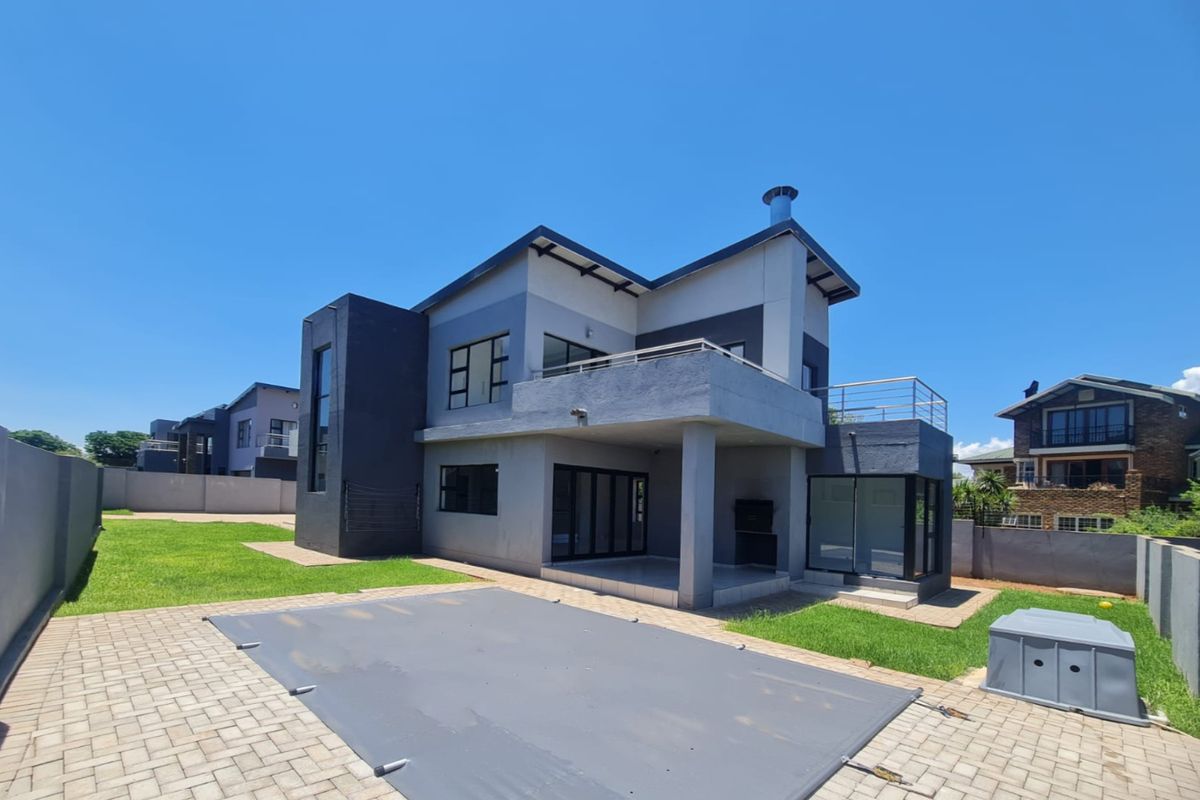 Rear view of a modern two-story house featuring a covered patio with built-in braai, an upper balcony, and a paved pool area.
