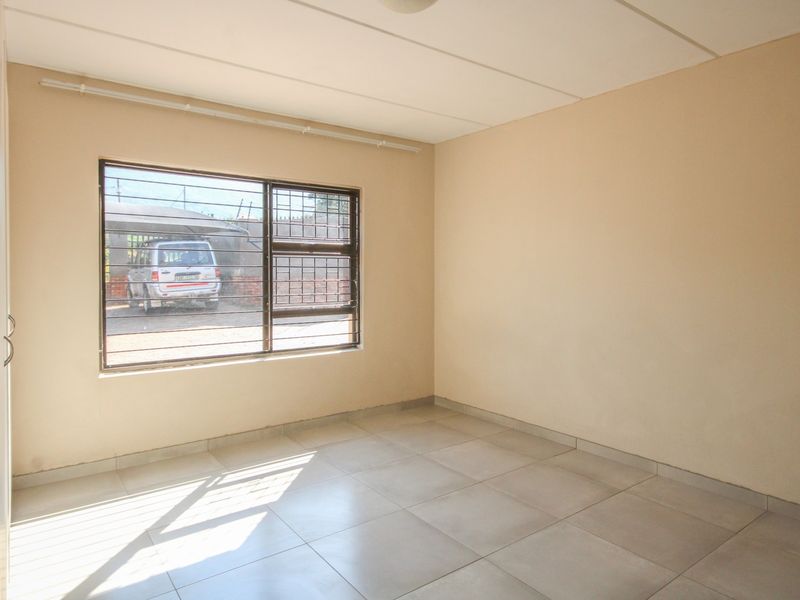 A bright, empty bedroom featuring neutral tiled flooring, built-in cupboard storage, and a window fitted with security bars.