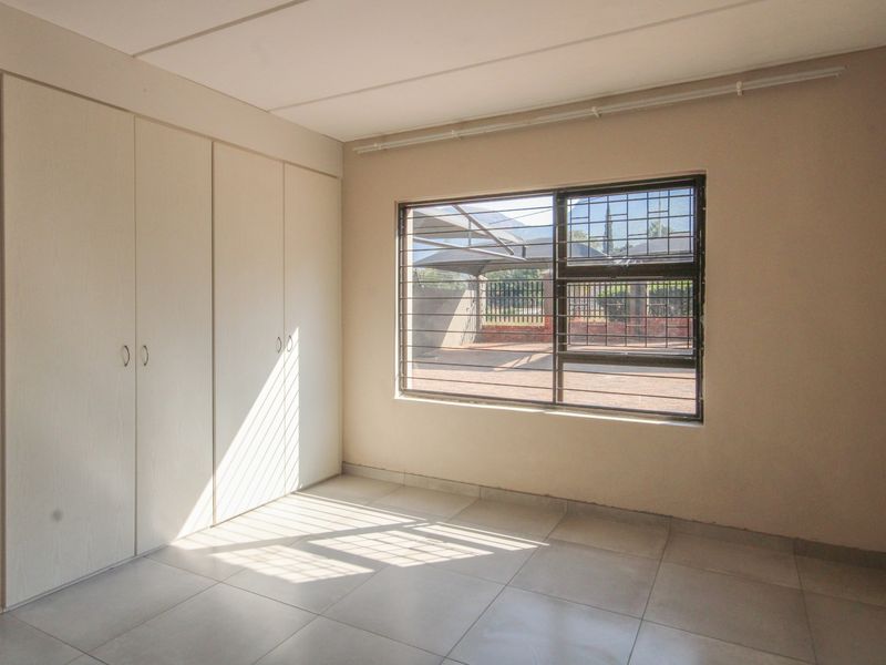 A bright, empty bedroom featuring neutral tiled flooring, built-in cupboards, and a window fitted with security bars.