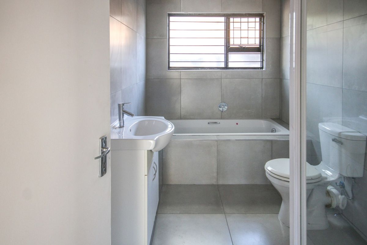 A clean, modern bathroom featuring neutral grey wall and floor tiles, a built-in bathtub, a pedestal basin, and a toilet.