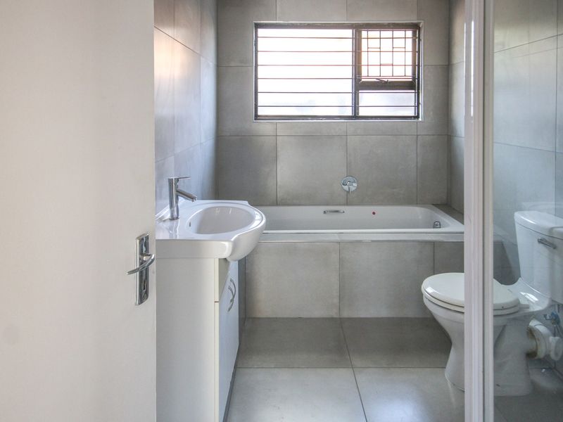 A clean, modern bathroom featuring neutral grey wall and floor tiles, a built-in bathtub, a pedestal basin, and a toilet.