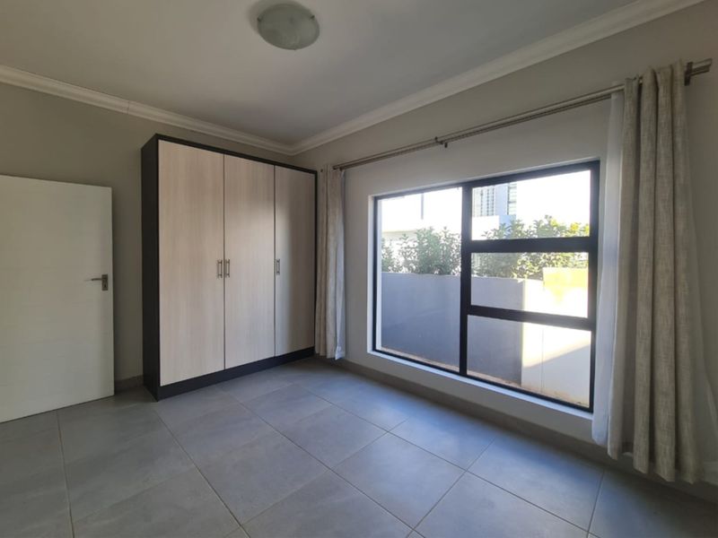 A bright, empty bedroom featuring neutral tiled flooring, built-in cupboards, and a large window with natural light.