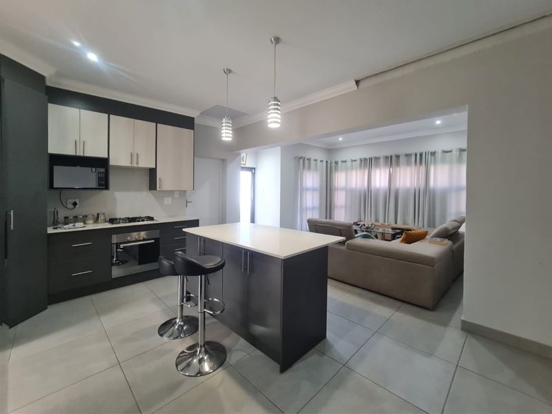 Modern open-plan kitchen and living area featuring a central island, sleek cabinetry, built-in appliances, and neutral tiled flooring throughout.