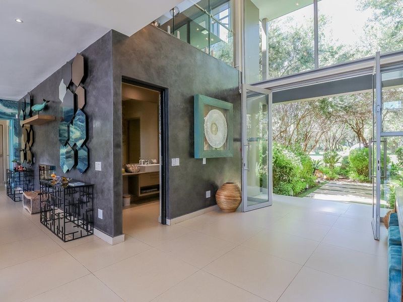 Modern living area featuring double volume ceilings, large format floor tiles, and expansive glass doors opening to a lush garden view.