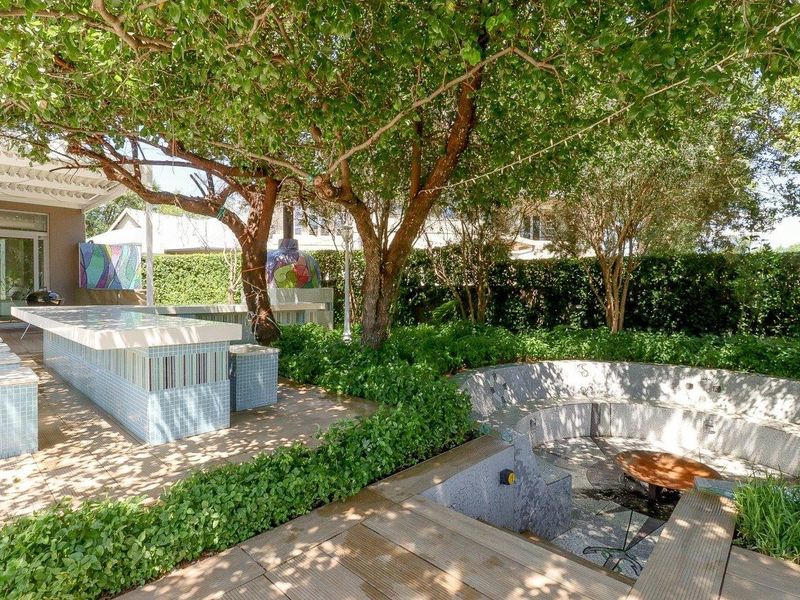 Outdoor entertainment area featuring a tiled bar, sunken seating, built-in pizza oven, and pergola, set within a lush landscaped garden.