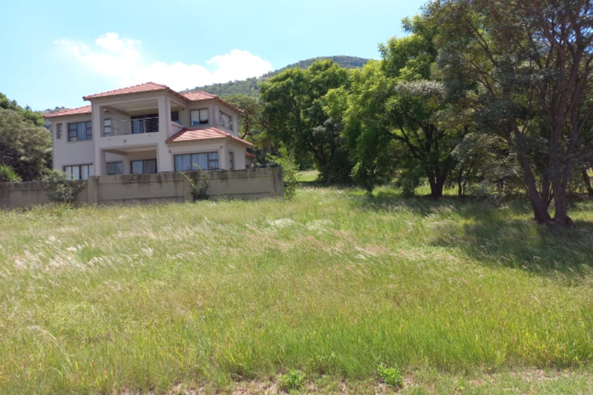 A view of a vacant residential plot featuring natural grass coverage, mature trees, and a neighboring double-story house with a tiled roof.