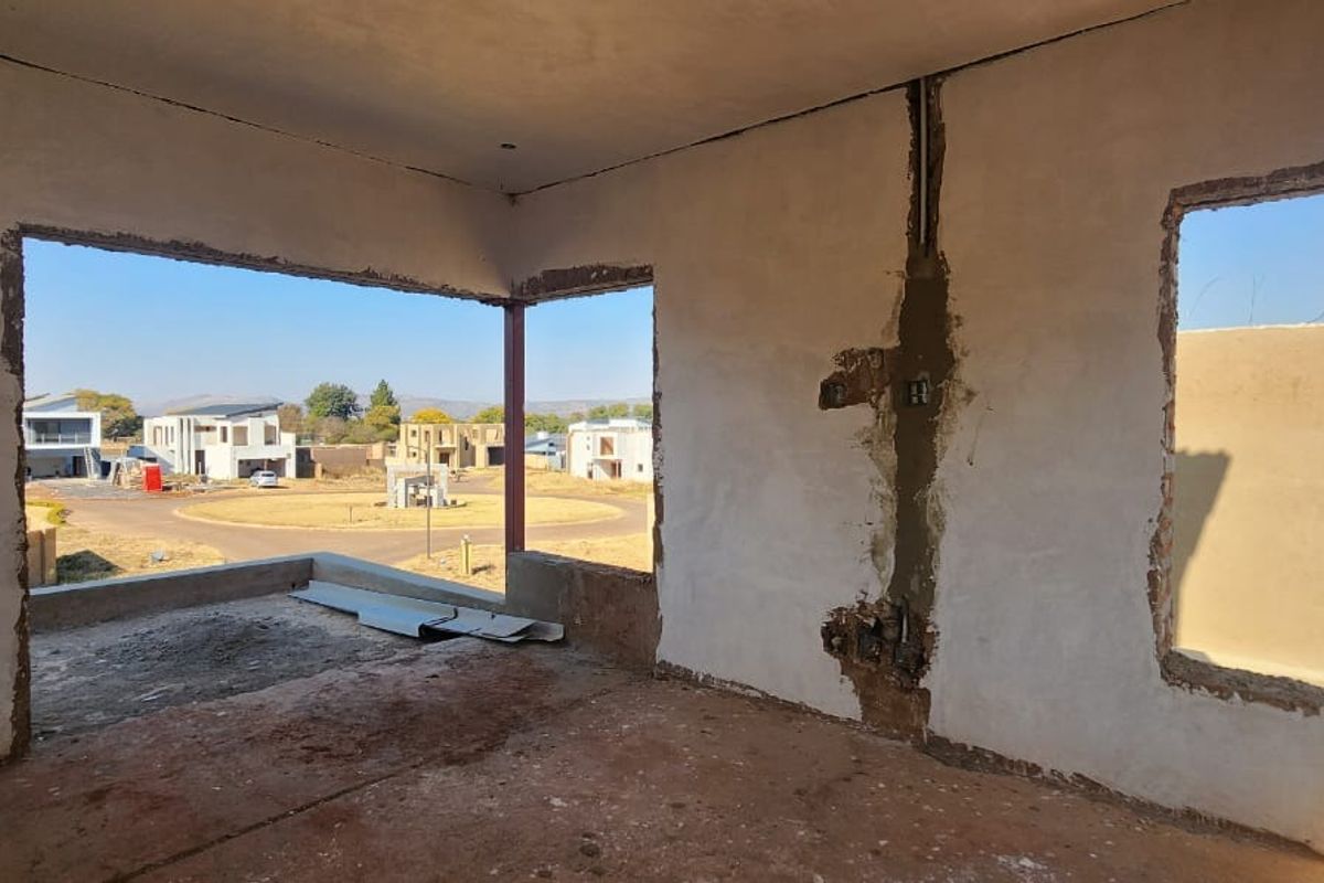 An interior view of a room under construction, featuring unfinished plastered walls, exposed electrical wiring, and large window openings with views of the surrounding estate.