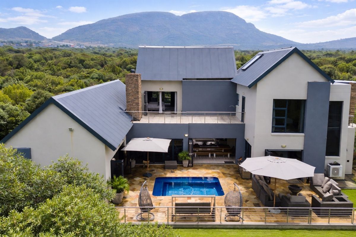 Aerial view of the rear exterior featuring a swimming pool, stone-paved patio, and a multi-level modern home with metal roofing and mountain views.
