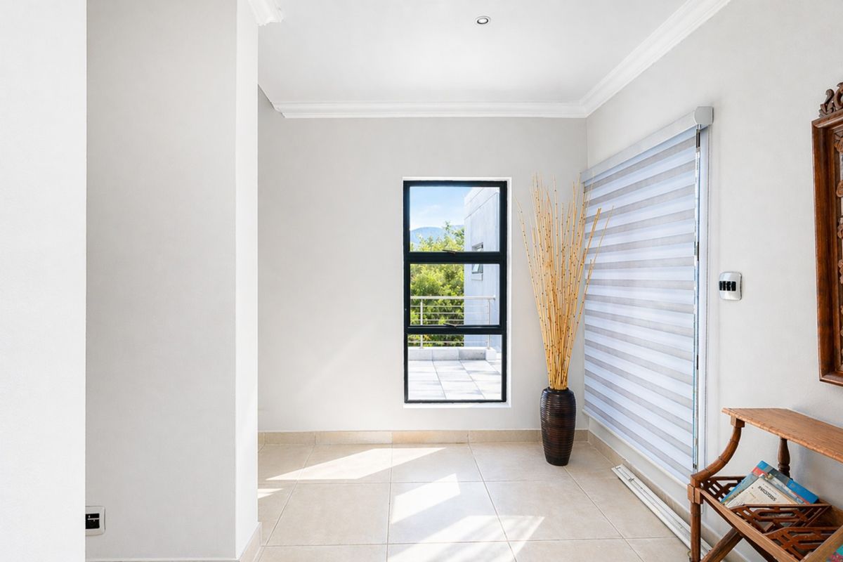 A bright, neutral-toned room featuring tiled flooring, modern window blinds, and a window providing natural light and access to an outdoor balcony area.